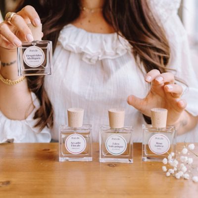 Cô gái cầm chai Hespérides Idylliques bên cạnh các dòng nước hoa thuần chay Poécile gồm Arcadie Florale, Eden Volcanique và Rose Lutèce trên bàn gỗ.