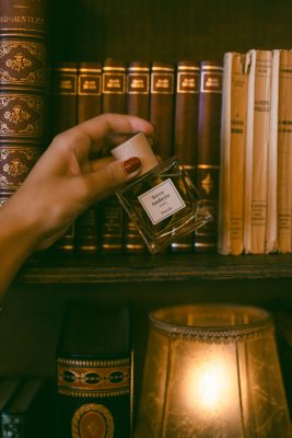 Bàn tay cầm chai nước hoa niche thuần chay Poécile Terre Ambrée Alsace nắp gỗ bên kệ sách cổ điển.