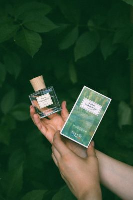 Đôi tay nâng chai và vỏ hộp nước hoa thuần chay Poécile Eden Volcanique Auvergne nắp gỗ sồi giữa tán lá xanh.