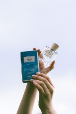 Đôi bàn tay nâng chai nước hoa thuần chay Poécile Bleu Asgard Bretagne và vỏ hộp xanh biển hướng lên nền trời khoáng đạt.