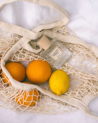 Chai nước hoa thuần chay Poécile Hespérides Idylliques Côte d'Azur nắp gỗ sồi đặt trong túi lưới cùng quả cam và chanh vàng tươi mát.