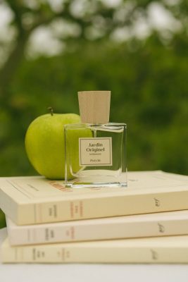 Chai nước hoa thuần chay Poécile Jardin Originel Normandie nắp gỗ đặt trên chồng sách cạnh quả táo xanh giòn.