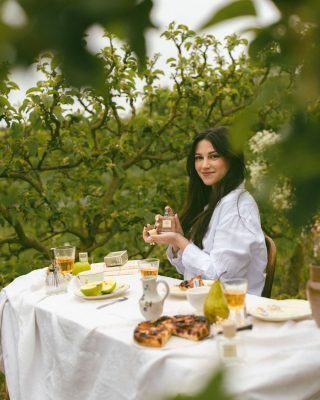 Cô gái mặc áo sơ mi trắng cầm chai nước hoa thuần chay Poécile Jardin Originel Normandie bên bàn tiệc trà dã ngoại giữa vườn táo xanh mát.   