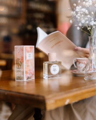 Chai nước hoa thuần chay Poécile Rose Lutèce Paris nắp gỗ và hộp đặt trên bàn gỗ quán cà phê cạnh tách trà, hoa baby trắng và người đọc sách.   