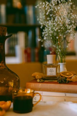 Chai nước hoa thuần chay Poécile Terre Ambrée Alsace đặt trên chồng sách cũ cùng gia vị khô, lát cam sấy và bình hoa baby trắng.