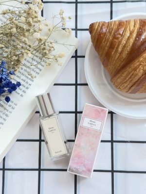 Chai nước hoa mini Poécile Rose Lutèce nắp bạc nhỏ gọn cùng vỏ hộp hồng bên bánh sừng bò croissant và trang sách mở.   