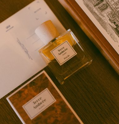 Chai nước hoa thuần chay Poécile Terre Ambrée Alsace nắp gỗ đặt trên tấm bưu thiếp cạnh cuốn sách bìa da cổ điển.   