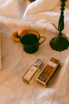 Chai nước hoa mini Poécile Terre Ambrée nắp bạc và vỏ hộp vân gỗ bên tách cà phê đen cùng chân nến cổ điển.