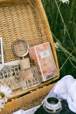 Chai nước hoa thuần chay Poécile Rose Lutèce Paris nắp gỗ đặt trong giỏ mây đan dã ngoại cùng kính lúp, bưu thiếp và máy ảnh cổ điển.   